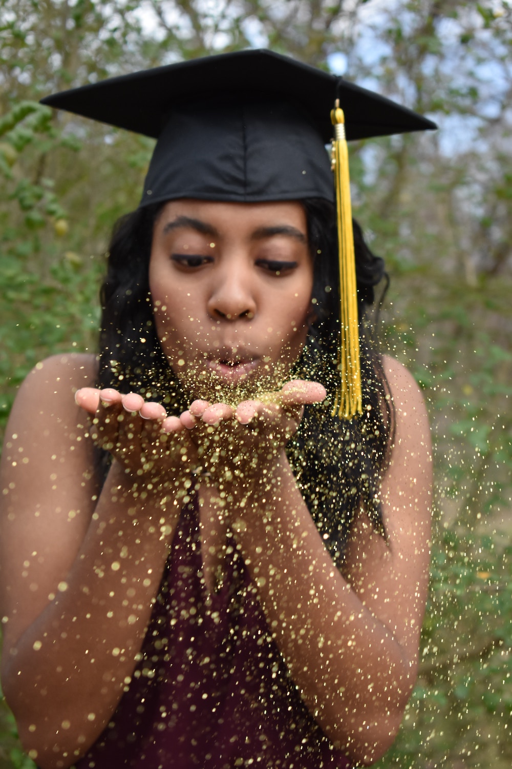 Girl wearing a graduation cap blowing on gold colored confetti.